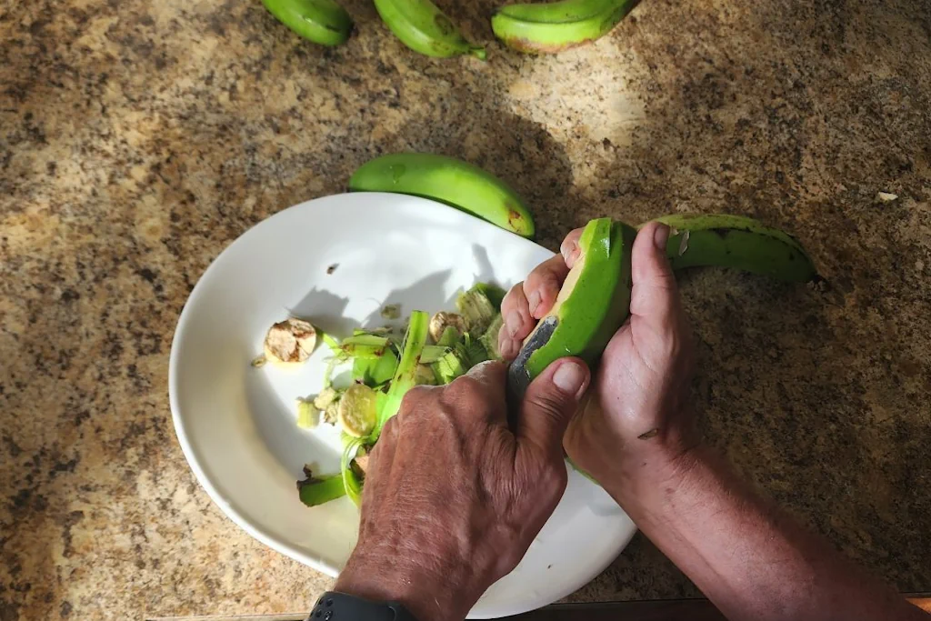 Peeling green bananas with a butter knife over a large bowl to catch the scraps.